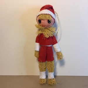 May include: A vintage Christmas bauble featuring a straw-haired doll wearing a red and white Santa suit. The doll has a smiling face with painted eyes and a gold string for hanging.
