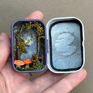 May include: A miniature diorama inside a small purple metal tin featuring two orange-capped mushrooms with white spots, two dark brown tree trunks with yellow foliage, and a moon illustration on the back wall. The lid shows a stippled moon design.