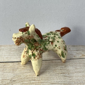 May include: A ceramic horse figurine with a cream-colored base, adorned with green and red speckled patterns. The mane and tail are a rich brown. The horse stands on four legs, showcasing a rustic, handcrafted aesthetic.