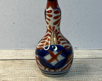May include: A small, white ceramic vase with a bulbous body and a narrow neck. The vase is decorated with a colorful pattern of red, blue, and white flowers and leaves.