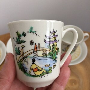 May include: White ceramic teacup with a colorful hand-painted design of a Japanese garden scene with a bridge, pagoda, and figures. The cup has a rounded shape and a handle.