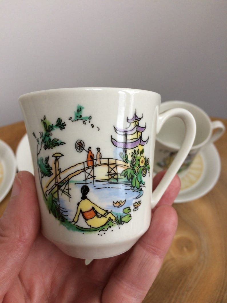May include: White ceramic teacup with a colorful hand-painted design of a Japanese garden scene with a bridge, pagoda, and figures. The cup has a rounded shape and a handle.