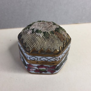 Könnte beinhalten: Eine kleine, sechseckige, dekorative Schachtel mit einem floralen Stoffdeckel. Die Schachtel ist aus Metall und hat ein weißes und blaues Emaille-Design mit goldenen Akzenten.