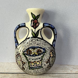 May include: A small white ceramic vase with blue and green floral designs and a mosaic pattern. The vase has two handles and a narrow neck.