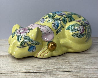 May include: A yellow ceramic cat figurine with a floral design. The cat is lying down with its eyes closed and has a gold bell on its ear.