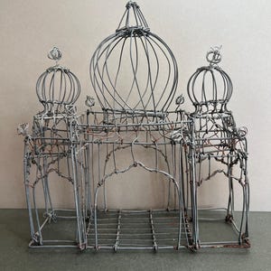 May include: A handcrafted wire sculpture of a building with three domes and arched entryways. The gray metal structure has a cage-like appearance, with intricate details and a touch of rust. The sculpture is a decorative piece.