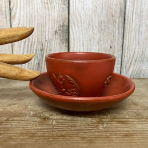 May include: A red clay teacup and saucer set with a floral design. The cup and saucer are both decorated with a raised floral pattern.