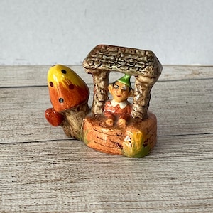 May include: A ceramic figurine of a gnome sitting in a mushroom house. The gnome is wearing a green hat and a red shirt. The mushroom house is brown and has a thatched roof.