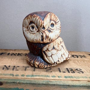 May include: A ceramic owl figurine featuring a textured, two-tone finish with dark brown and cream-colored glaze. The owl has deeply carved facial features, large circular eyes, and detailed wing patterns, showcasing a rustic, hand-crafted appearance.
