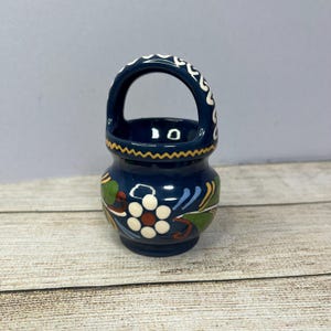 May include: A small blue ceramic pot with a white handle and floral design. The pot has a yellow rim and a white flower with a red centre.