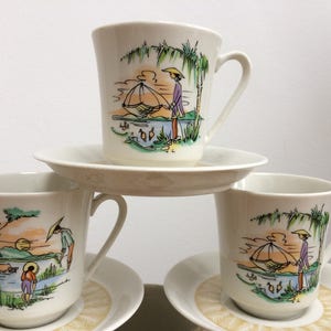 May include: Three white teacups with saucers, each featuring a colorful illustration of a person in a boat with a fishing net, surrounded by palm trees and water. The cups and saucers have a delicate, vintage design.