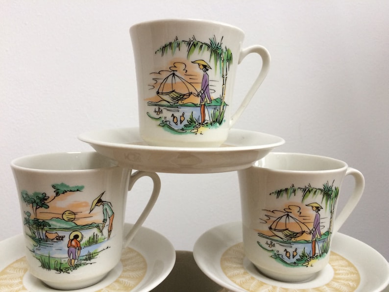 May include: Three white teacups with saucers, each featuring a colorful illustration of a person in a boat with a fishing net, surrounded by palm trees and water. The cups and saucers have a delicate, vintage design.