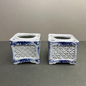 May include: Two square, white ceramic containers with blue floral designs. Each container has a lattice pattern on the sides and a circular opening at the top. The containers are likely for decorative purposes.
