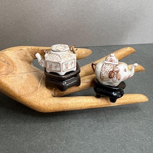 May include: Two miniature porcelain teapots, one square and one elephant-shaped, sit on black stands. The square teapot has a floral design with gold accents. The elephant teapot features a brown and white pattern. Both are displayed on a wooden surface.