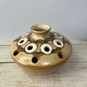May include: A small, round, ceramic vase with a brown and white design. The vase has multiple holes around the top for holding flowers.