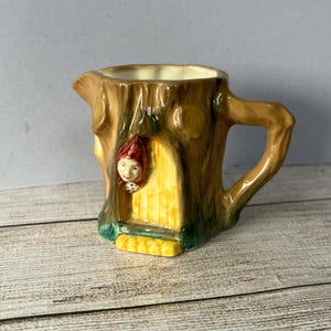 May include: A ceramic creamer in the shape of a tree trunk, with a small door and a face wearing a red hood. The creamer has a brown exterior with yellow and green details, and a handle on the side.