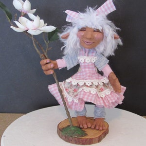May include: A handmade doll with white hair, wearing a pink and white gingham dress, holding a branch with white flowers. The doll is standing on a wooden base.