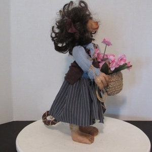 May include: A handmade doll with brown hair, a blue shirt, a brown vest, and a striped skirt. The doll is holding a basket of pink flowers.