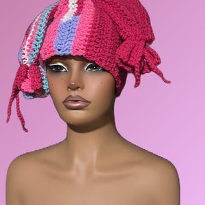 Puede incluir: Un sombrero de ganchillo hecho a mano en tonos rosa, azul y blanco. El sombrero presenta un diseño a rayas en la parte delantera y una sección rosa lisa en la parte trasera con borlas colgantes. El sombrero se muestra en una cabeza de maniquí sobre un fondo rosa.