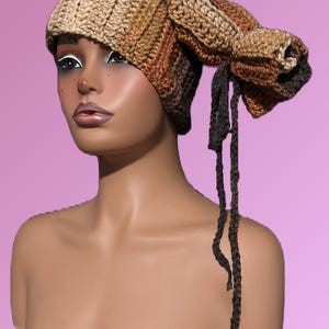 Puede incluir: Un sombrero de ganchillo hecho a mano en tonos beige, marrón y negro. El sombrero presenta un diseño único y alargado con una parte superior puntiaguda y una larga corbata trenzada. El sombrero se exhibe en un maniquí sobre un fondo rosa.