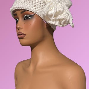 Puede incluir: Un sombrero blanco de ganchillo con un diseño único. El sombrero está hecho de un material tejido texturizado y presenta un acento decorativo y con volantes en el lateral. El sombrero se exhibe en un maniquí con un fondo rosa.