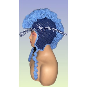 May include: A blue and navy blue crocheted hat with a unique design. The hat has a large, ruffled crown and long, curly tails. The hat is worn on a mannequin's head. The text "@phoenix_the_entrepreneur" is visible on the hat.