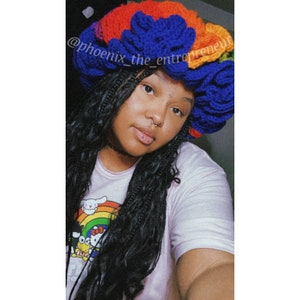 May include: A person wearing a rainbow crocheted flower crown with blue and orange accents. The crown is made of yarn and has a fluffy texture. The person is wearing a pink shirt with a cartoon graphic and has long black hair.
