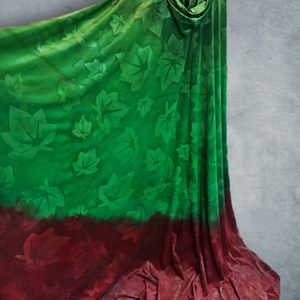 Pode incluir: Uma capa com capuz apresentando um design em degradê que transita de um verde vibrante no topo para um bordô profundo na base. O tecido é adornado com um padrão repetitivo de formas de folhas em relevo por toda a peça.