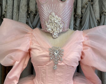 May include: A light pink dress featuring voluminous sheer puff sleeves, a structured bodice, and a large silver-toned brooch with clear crystals. Above the dress sits a matching light pink crown adorned with silver glitter, pearls, and white floral accents.