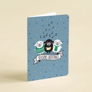 Könnte beinhalten: Eine blaue Karte mit weißem Rand und einem festlichen Design mit drei singenden Schafen. Die Schafe tragen Schals und das schwarze Schaf hält ein Notenbuch. Der Text auf der Karte lautet "Seasons Bleatings".