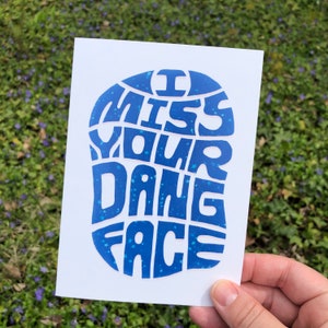 May include: A white greeting card featuring the phrase "I MISS YOUR DANG FACE" in a blue, decorative font. The text is contained within a blue, rounded shape with star details. The card is held against a backdrop of green grass and small purple flowers.