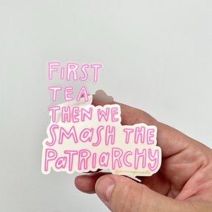 以下が含まれることがあります： 「First Tea Then We Smash The Patriarchy」というテキストが書かれたピンクのステッカー。ステッカーの下には小さな著作権記号と「@mamaparade」というテキストがあります。
