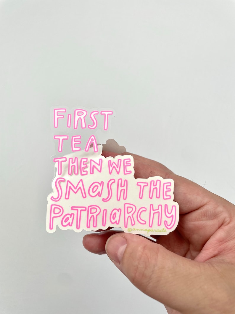 以下が含まれることがあります： 「First Tea Then We Smash The Patriarchy」というテキストが書かれたピンクのステッカー。ステッカーの下には小さな著作権記号と「@mamaparade」というテキストがあります。