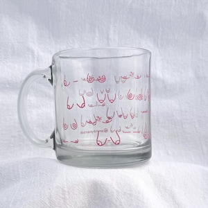May include: A clear glass mug with a pink outline of various breasts printed on the side. The mug has a handle and is sitting on a white surface.