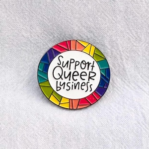 May include: A round enamel pin with a rainbow design and the text "Support Queer Business" in black lettering.