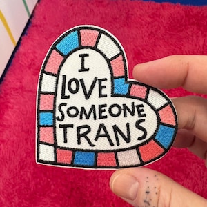 Op de afbeelding: Witte, hartvormige geborduurde patch met een roze, blauwe en witte rand. De patch zegt "Ik hou van iemand die trans is" in zwarte tekst.