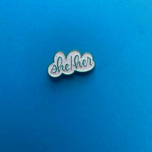 May include: A white cloud-shaped enamel pin with green outlining and glitter. The pin says "she/her" in cursive.