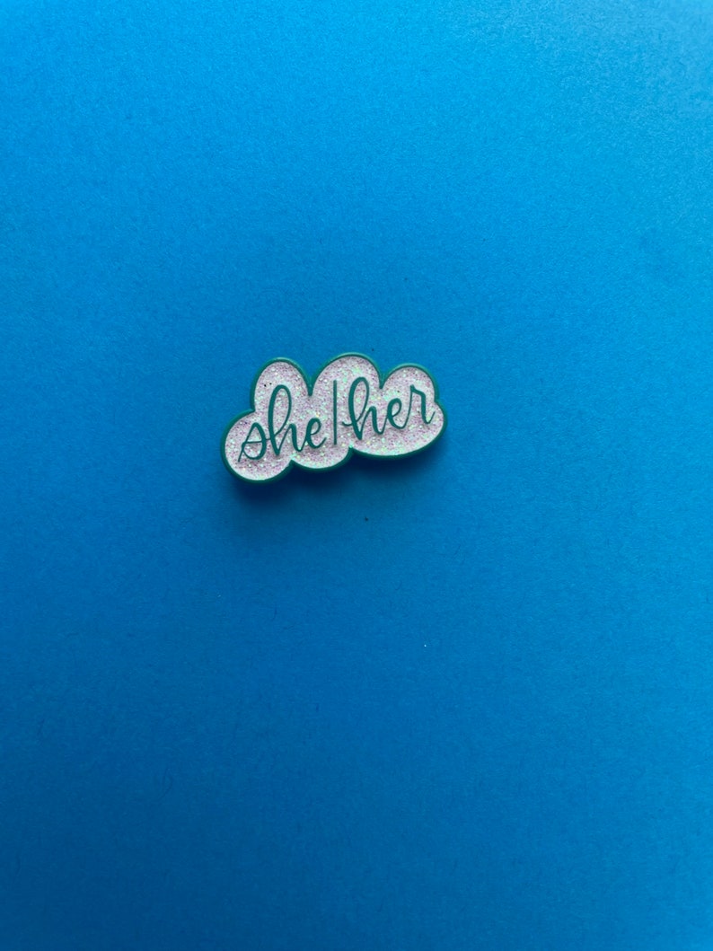 May include: A white cloud-shaped enamel pin with green outlining and glitter. The pin says "she/her" in cursive.