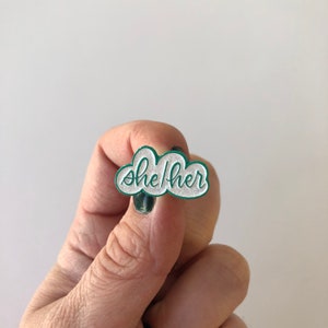 May include: A green enamel pin shaped like a cloud with the words "she/her" written in white glitter.