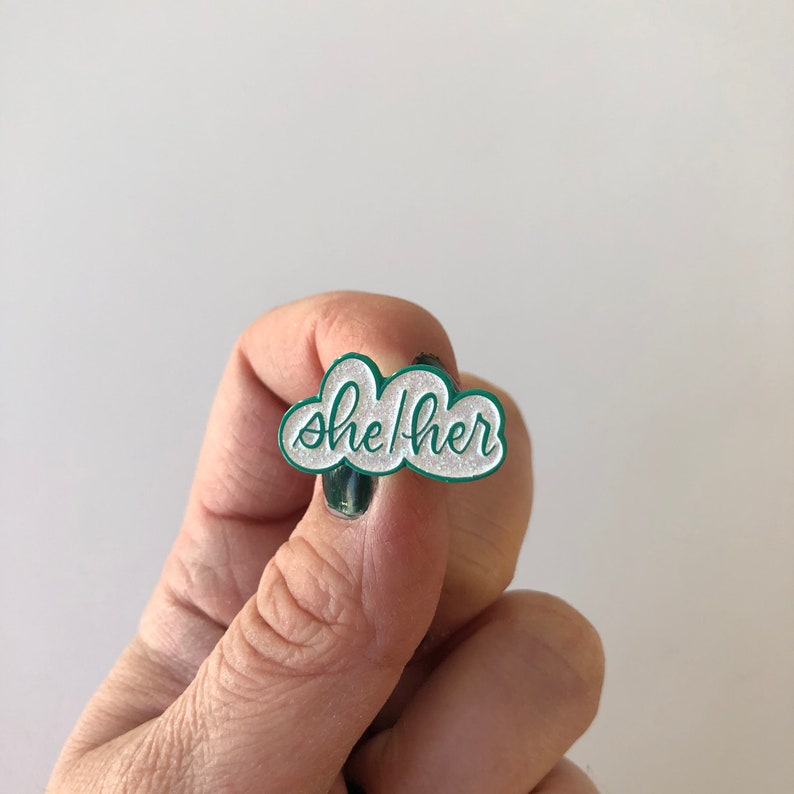 May include: A green enamel pin shaped like a cloud with the words "she/her" written in white glitter.