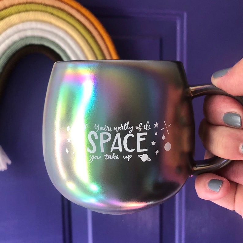 May include: A iridescent gray mug with a white inscription that reads "You're worthy of the space you take up". The mug has a handle and is decorated with small stars and a planet.
