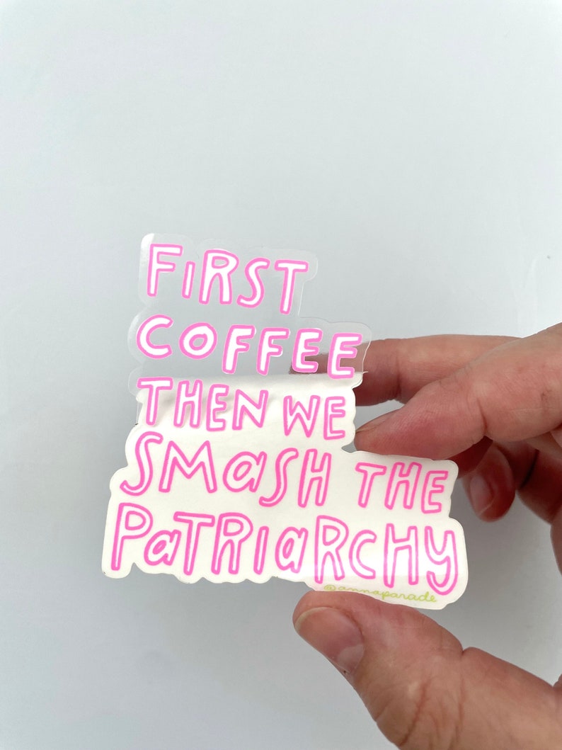 以下が含まれることがあります： 「First Coffee Then We Smash The Patriarchy」というテキストが書かれたピンクのステッカーで、下部に「@mamaparade」という小さな緑色のテキストがあります。