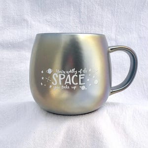 May include: A silver iridescent mug with a black handle. The mug has a white inscription that reads "You're worthy of the space you take up" with a small planet and star graphic.