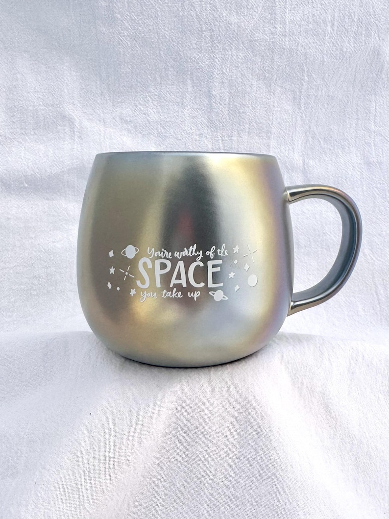 May include: A silver iridescent mug with a black handle. The mug has a white inscription that reads "You're worthy of the space you take up" with a small planet and star graphic.