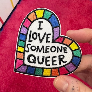 Puede incluir: Un parche blanco en forma de corazón con un borde de arcoíris. El parche dice "Amo a alguien queer" en texto negro.