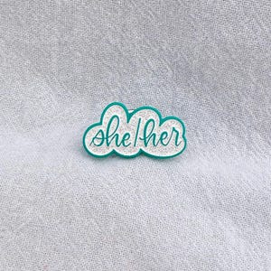 May include: A teal blue enamel pin in the shape of a cloud with the text "she/her" in white glitter.