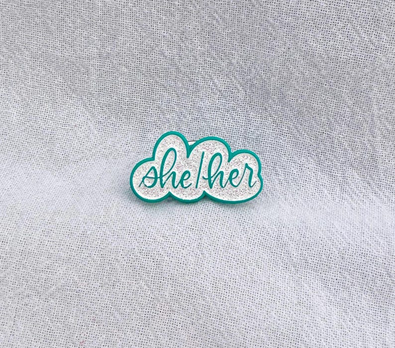 May include: A teal blue enamel pin in the shape of a cloud with the text "she/her" in white glitter.