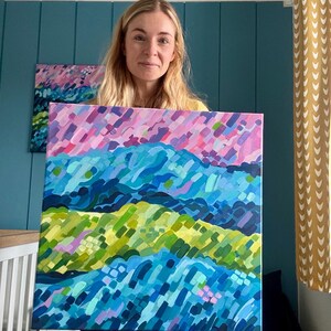 Puede incluir: Una persona sostiene una colorida pintura abstracta de un paisaje. La obra de arte presenta capas de pinceladas azules, verdes y rosas, creando un efecto texturizado. Una pintura m&aacute;s peque&ntilde;a con colores similares es visible en el fondo.