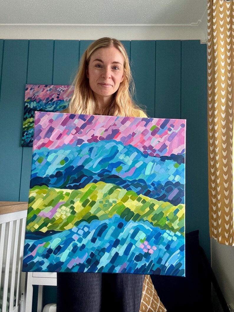 Puede incluir: Una persona sostiene una colorida pintura abstracta de un paisaje. La obra de arte presenta capas de pinceladas azules, verdes y rosas, creando un efecto texturizado. Una pintura m&aacute;s peque&ntilde;a con colores similares es visible en el fondo.