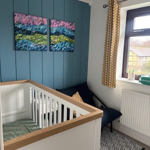 Puede incluir: Un cuarto de beb&eacute; con una cuna blanca con detalles de madera, una pared decorativa azul y dos pinturas abstractas de paisajes. Una silla azul marino con un coj&iacute;n estampado se encuentra junto a una ventana con cortinas amarillas. Una alfombra geom&eacute;trica en blanco y negro est&aacute; en el suelo.